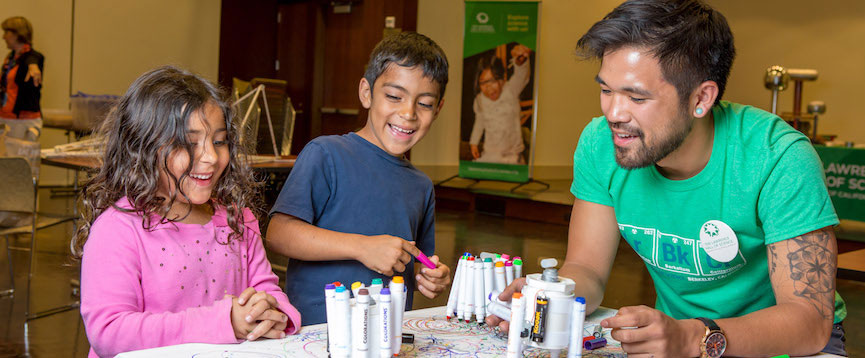 Two children and an adult are siting at a drawing table station and working on their project.